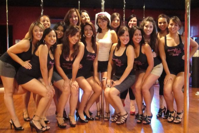 Bachelorette party group of smiling women in tank tops, shorts and heels posing around pole-dance poles in a dance studio