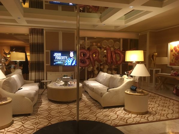 Elegant hotel-suite lounge with two cream sofas facing a round coffee table with drinks, a chrome dance pole in the center, warm lamps, and rose-gold bridal balloons against a gold-disc backdrop, TV showing a musician promo.