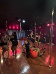 Energetic pole-dance fitness class in a dim studio with red accent lights and hardwood floor — a group of women in black (one in red) striking poses on gold poles.
