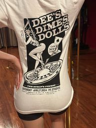 Back view of person wearing a white graphic tee showing a pinup dancer on a roulette wheel with playing cards and Las Vegas event text, standing by a brass pole on a shiny wooden floor.