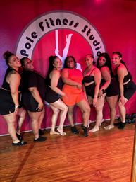 Seven women in workout clothes smiling and posing together in front of a bright pink pole fitness studio logo wall on a wood floor — playful group photo from a studio session.