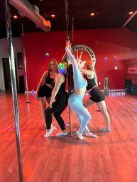 Four women in activewear posing around a dance pole in a red-walled pole fitness studio with wood floors and colorful disco lights.