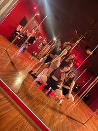 Five women taking a mirror selfie in a red-lit pole dance studio with hardwood floors, chrome poles and a disco ball, energetic pole fitness class vibe.