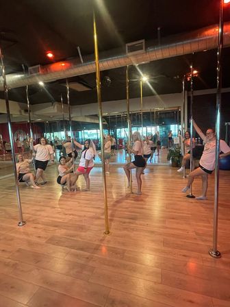 Group of women in a pole fitness class posing on shiny poles in a mirrored dance studio with hardwood floors and exposed ductwork, smiling and striking playful poses.