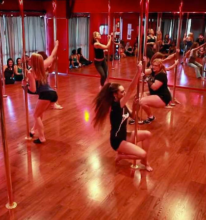 Energetic pole dance fitness class with women practicing pole moves in a red mirrored studio on polished hardwood floors, others seated along the wall.