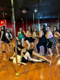 Group photo of women in workout clothes posing around chrome dance poles in a pole dance fitness studio with wood floors, red walls and a disco ball.