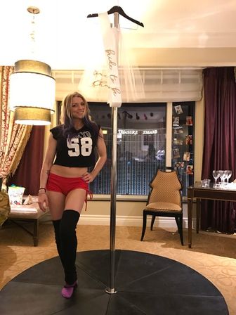 Playful bachelorette scene in a hotel room: person in a black '58' crop top, red shorts and thigh-high socks posing by a dance pole with a "bride to be" sash hanging above and city lights visible through the window.