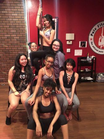 Seven women in athletic wear smiling and posing inside a pole fitness studio — one climbs a chrome pole while others sit on chairs against a red wall and wood floor.