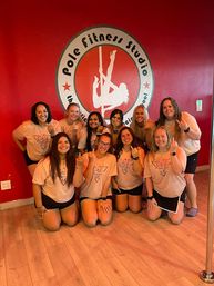 Smiling group of women in matching peach T-shirts posing inside a pole fitness studio against a red wall with a pole logo, playfully showing middle-finger gestures.