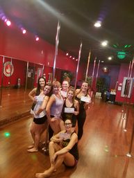 Seven women in a pole fitness studio posing with certificates around chrome poles on a wooden floor with red walls and mirrors, celebrating a pole-dance class completion