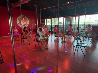 Group of women in a pole fitness studio taking a chair-based dance class under colorful disco lights on a wood floor, one participant wearing a bridal veil.