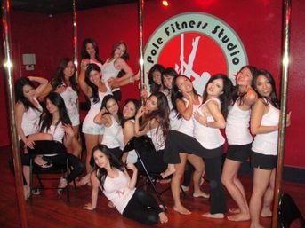 Group of women in matching white tank tops posing and smiling around chrome poles in a pole fitness studio with a red wall and circular logo.