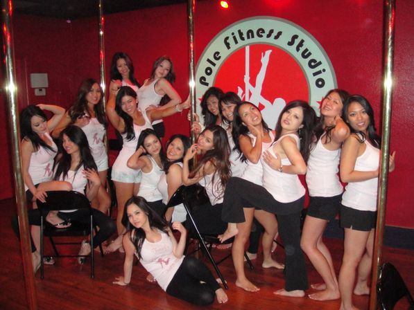 Group of women in matching white tank tops posing and smiling around chrome poles in a pole fitness studio with a red wall and circular logo.