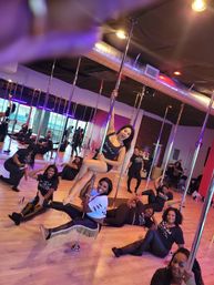 Group of women laughing and posing in a lively pole fitness class inside a mirrored dance studio with hardwood floors, metal poles and colorful lighting.