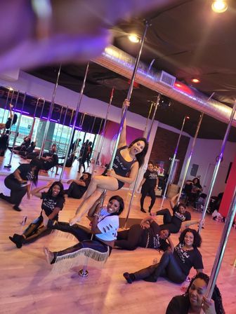 Group of women laughing and posing in a lively pole fitness class inside a mirrored dance studio with hardwood floors, metal poles and colorful lighting.