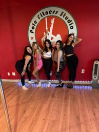 Five women in activewear pose and smile against a red wall with a pole-fitness logo, wooden floors, a pole and LED strip lights — upbeat group workout vibe.