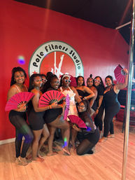 Group of women in black workout outfits and one in white posing barefoot inside a pole fitness studio against a red wall with a circular logo, smiling and holding bright pink folding fans around a chrome pole.