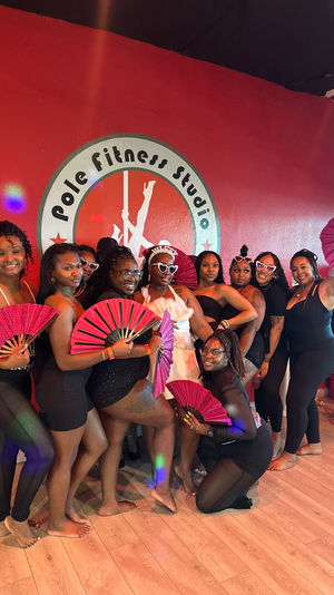 Group of women in black workout outfits and one in white posing barefoot inside a pole fitness studio against a red wall with a circular logo, smiling and holding bright pink folding fans around a chrome pole.