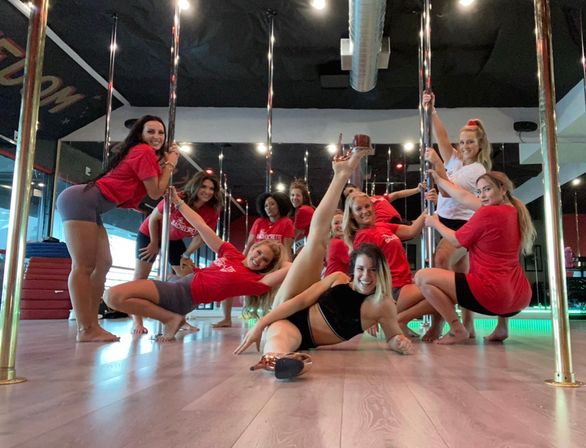 Group of women in a pole fitness class at a dance studio — smiling friends in red shirts and activewear pose around chrome poles, one woman on the floor with a high leg split, mirrored walls and LED lighting in the background.