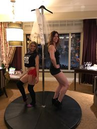 Two women posing back-to-back on a portable dance pole in a hotel suite for a bachelorette celebration, veil sash hanging above and city skyline visible through the window.