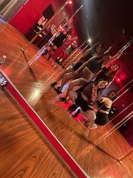 Energetic group of women posing for a mirror selfie during a pole fitness class in a red-lit studio with hardwood floors and metal poles.