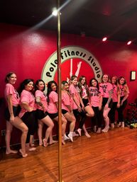 A group of women in matching pink shirts striking playful poses barefoot beside a brass pole in a bright red pole-fitness studio with a circular wall logo and wooden floors.
