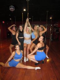 Seven women in athletic wear posing around a brass pole in a pole-fitness dance studio with wood floors and a disco ball