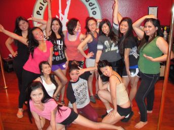 Group of women posing playfully in a pole dance fitness class inside a red-walled studio with wooden floor and chrome poles