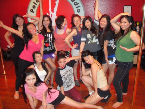 Group of women posing playfully in a pole dance fitness class inside a red-walled studio with wooden floor and chrome poles