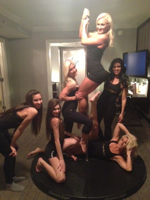 Six women in black outfits posing on and around a portable dance pole on a round stage inside a hotel suite — playful girls' night pole-dancing party.