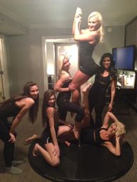 Six women in black outfits posing on and around a portable dance pole on a round stage inside a hotel suite — playful girls' night pole-dancing party.
