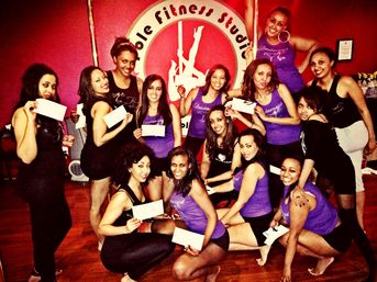 Smiling group of women posing in a pole-fitness studio, many wearing purple tank tops and black shorts and holding certificates — celebratory pole dance class photo.
