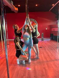 Four women in athletic wear striking playful poses around a chrome pole in a red-walled pole fitness studio with wood floors and colorful lights, group dance/workout session.