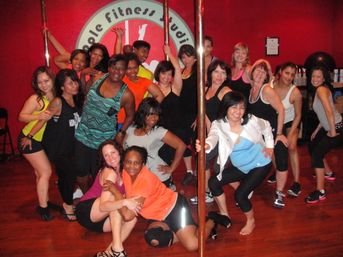 Group of diverse women smiling and posing in a bright red indoor pole-fitness studio around brass poles on a wooden floor, playful group fitness class vibe.