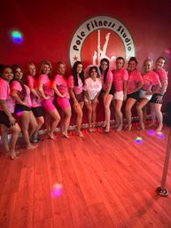 Smiling group in matching pink shirts posing in a pole fitness studio with red walls and wooden floor, bride-to-be in white and veil at the center next to a dance pole.