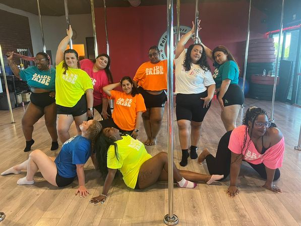 Group of women in colorful "Bride Tribe" T-shirts posing around poles in a bright pole-dancing studio for a playful bachelorette class