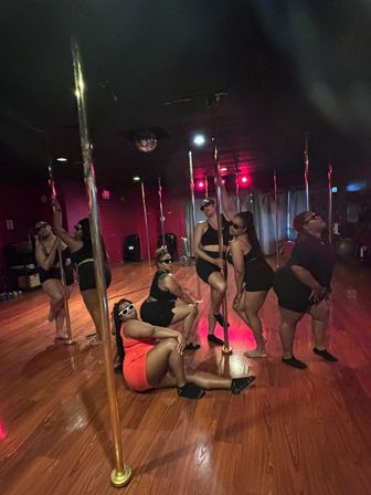 Group of women in athletic wear and sunglasses posing around brass poles in a red-lit pole fitness studio with polished hardwood floors.