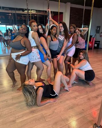 Playful group of women in activewear posing around a pole in a bright dance studio with mirrors and wood floors — pole fitness class vibe.