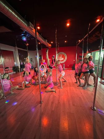 Smiling group of women in pink shirts practicing and posing with chrome poles in a red-lit pole dance fitness studio with wood floors