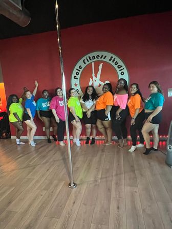Smiling group of women in bright neon shirts posing in a pole fitness studio, lined up against a red wall with a circular studio logo and a chrome dance pole on a light wood floor.