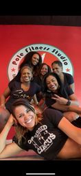 Six smiling women posing inside a pole-fitness studio, one reclining in front and five grouped behind against a red wall with a round studio logo; casual dark workout shirts and a fun, relaxed group vibe.