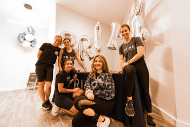 Smiling team of five female fitness instructors in athletic wear posing at a bright boutique indoor cycling studio welcome desk with silver balloons and celebratory decor.