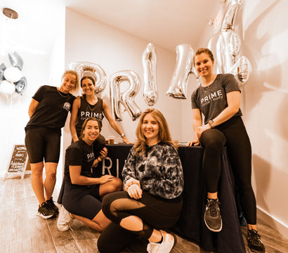 Smiling team of five female fitness instructors in athletic wear posing at a bright boutique indoor cycling studio welcome desk with silver balloons and celebratory decor.