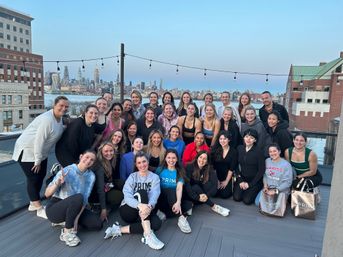 Smiling group of women on a New York City rooftop terrace at dusk after a group fitness class, with the Manhattan skyline and river in the background.