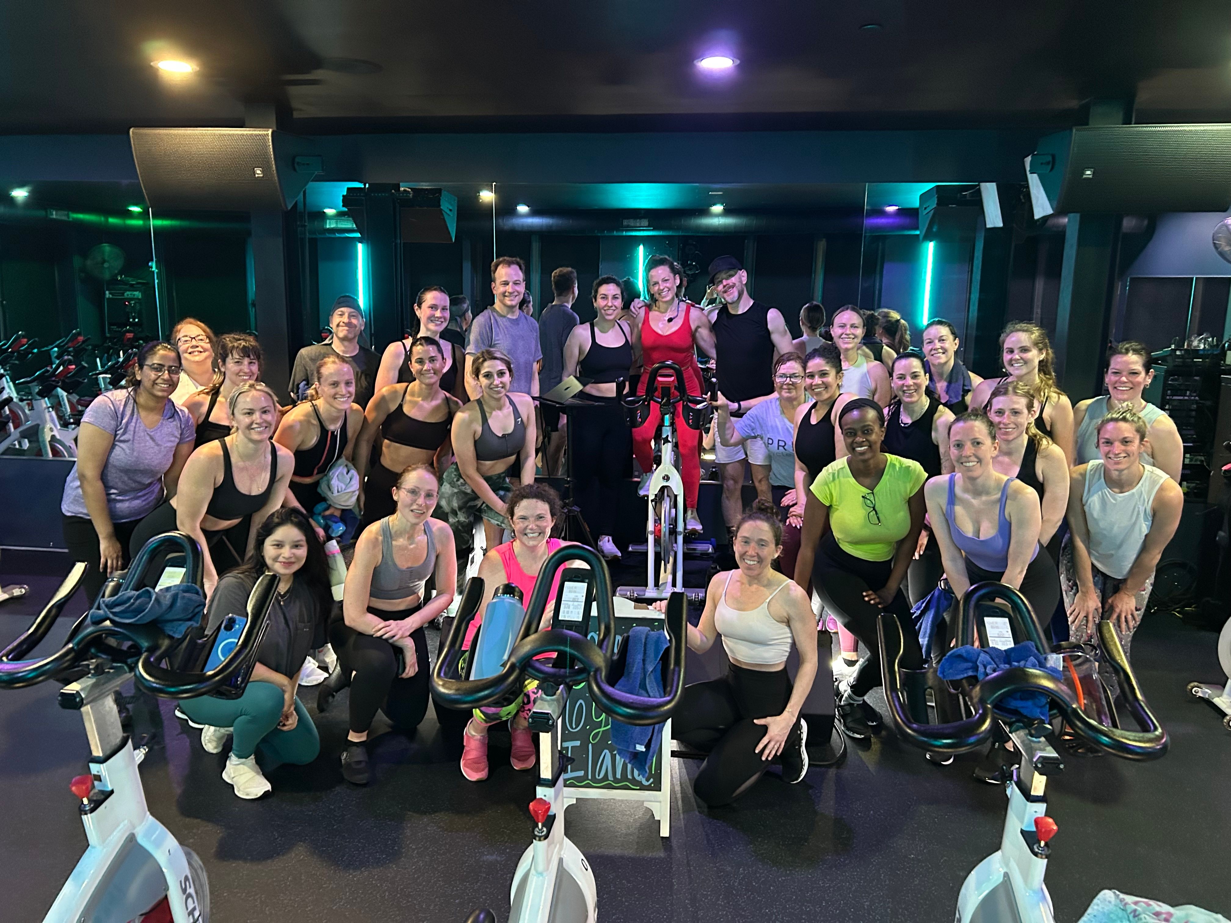 Cheerful group spin class posing with stationary bikes in a neon-lit indoor cycling studio after a workout, diverse riders smiling for a post-class group photo.