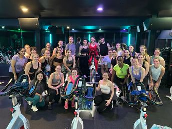 Cheerful group spin class posing with stationary bikes in a neon-lit indoor cycling studio after a workout, diverse riders smiling for a post-class group photo.