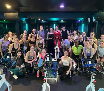 Cheerful group spin class posing with stationary bikes in a neon-lit indoor cycling studio after a workout, diverse riders smiling for a post-class group photo.