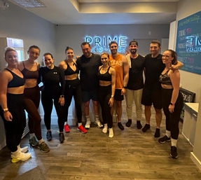 Cheerful group of eleven adults in athletic wear posing after a spin class inside an indoor cycling and fitness studio with hardwood floors and neon signage in the background
