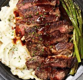 Juicy sliced medium-rare steak drizzled with glossy brown sauce, served with creamy mashed potatoes and grilled asparagus on a black plate