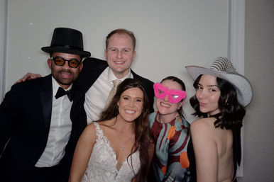 Smiling group of five in a wedding photo booth: woman in a lace white dress, two men in tux/suit (one wearing a top hat), a guest with pink fuzzy heart-shaped glasses, and a guest in a silver cowboy hat posing against a plain backdrop.